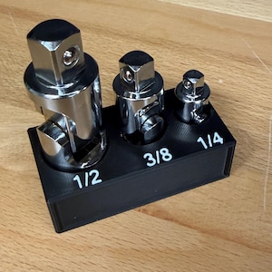 May include: A black plastic storage block holding three chrome universal joint socket adapters. The block features white printed labels for 1/2, 3/8, and 1/4 inch drive sizes, with each adapter seated in its corresponding circular slot on the base.