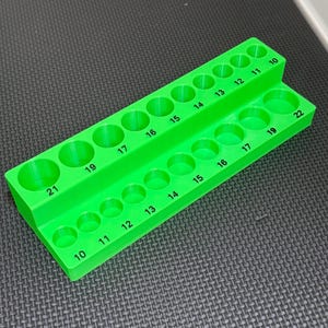 May include: A bright green, two-tiered organizer with numbered holes. The top tier has holes labeled 10 through 17, and the bottom tier has holes labeled 10 through 22. The organizer is made of a solid, opaque material and sits on a textured surface.