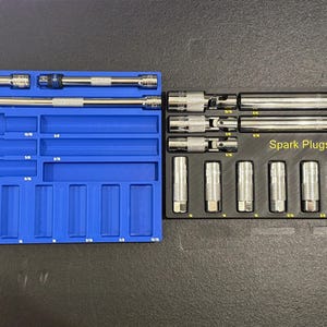 May include: Two automotive tool organizers. A blue organizer holds various tools, while a black organizer displays spark plugs and the text "Spark Plugs". The tools are silver and the organizers are on a dark surface.