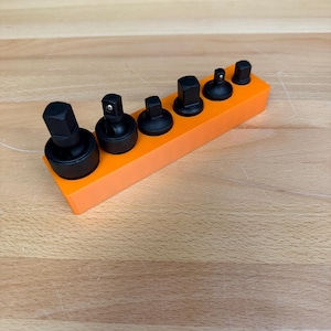 May include: An orange tool holder with seven black socket wrench attachments. The sockets are arranged in a row, varying in size and shape. The holder is rectangular and sits on a light-colored wooden surface.