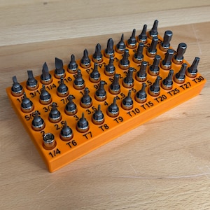 May include: A rectangular orange plastic organizer tray holding a large collection of metal screwdriver bits. The tray features printed labels for each bit, including sizes like T6, T7, T8, T9, T10, T15, T20, T25, T27, T30, 1/4, 3/16, and 5/64.