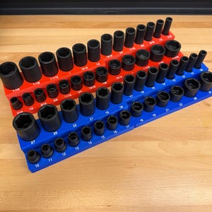 May include: Three socket organizers in red and blue, holding a variety of black sockets. The sockets are arranged by size, with measurements like 1/2, 5/8, and 3/4 inches visible. The organizers sit on a wooden surface.