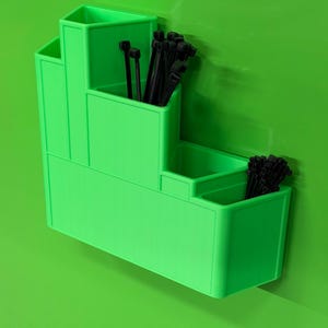 May include: A bright lime green tiered wall-mounted organizer featuring four distinct compartments of varying heights. The organizer is filled with multiple black plastic zip ties, which are visible protruding from the top of the storage sections.