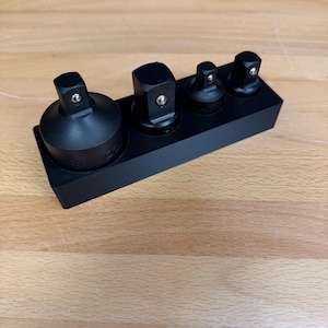 May include: A set of four black metal socket adapters arranged in a row on a rectangular black base. Each adapter features a square drive head with a small silver ball bearing mechanism for secure attachment, varying in size from largest to smallest.