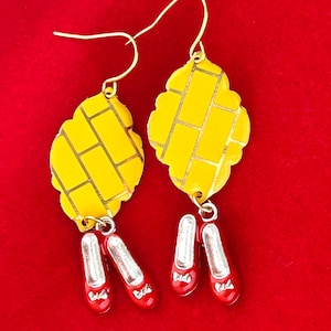 May include: A pair of gold-tone earrings featuring yellow brick road pendants and dangling red shoe charms. The yellow pendants have a brick pattern. The red shoes have silver accents and bows. The earrings hang on gold-tone hooks.