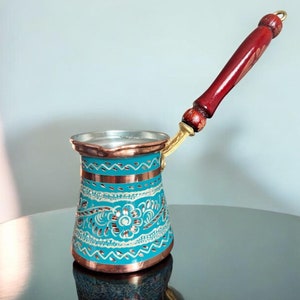 Puede incluir: Una cafetera turca tradicional con un diseño azul y cobre y un mango de madera. La cafetera está decorada con intrincados patrones florales.