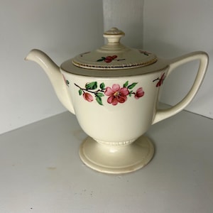 May include: A vintage cream-colored teapot with a floral design. The teapot has a lid with a gold trim and a base with a pedestal design. The flowers are pink and green.