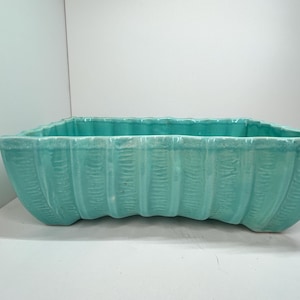 以下が含まれることがあります： 長方形のターコイズ色のセラミックプランター。リブ模様のデザインで、上端は波型、ベースは曲線的です。ターコイズ色はプランター全体に均一に施されています。