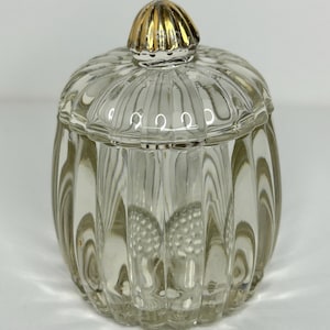 Könnte beinhalten: Ein klares Glasgefäß mit Deckel. Das Gefäß hat ein geripptes Design und einen goldfarbenen Knopf oben. Das Gefäß ist leer und steht auf einer weißen Oberfläche. Das Gefäß dient wahrscheinlich zur Aufbewahrung kleiner Gegenstände.