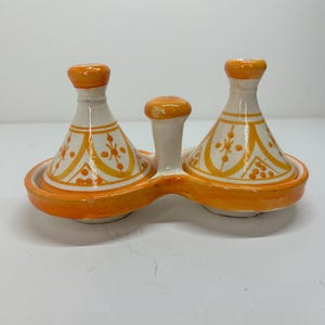 Puede incluir: Un servidor de condimentos de cerámica con dos tapas cónicas y un asa central. El servidor es blanco con detalles en naranja y patrones decorativos. La base y las tapas son naranjas, con una estética marroquí o de Oriente Medio.