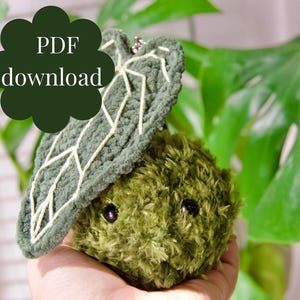 May include: A handmade green plush creature with a leaf-shaped hat, featuring black eyes and a fluffy texture. The leaf has white stitching. The text "PDF download" is in a dark green bubble.