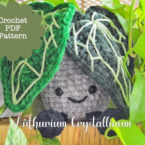 Puede incluir: Una planta de Anthurium Crystallinum tejida a ganchillo con una cara sonriente, cuerpo gris y hojas verdes oscuras. Las hojas tienen detalles de venas verde claro. El texto "Crochet PDF Pattern" está en una burbuja verde, y el texto "Anthurium Crystallinum" debajo.