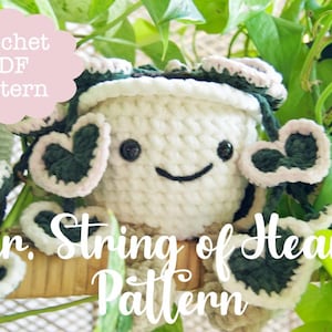 Pode incluir: Um vaso de plantas de crochê branco com um rosto sorridente e folhas em forma de coração, com acabamento em verde escuro e rosa claro. O texto "Van. String of Hearts Pattern" é visível.