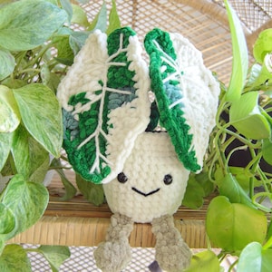 May include: A handmade, smiling crochet plant with large, white and green leaves. The plant has a cream-colored pot and gray, stubby legs. The leaves are detailed with green and blue accents. The plant is surrounded by green foliage.