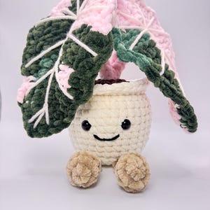 May include: A crocheted plant with a smiling face. The leaves are pink and green, with white veins. The pot is off-white, and the plant has two small, tan pom-pom feet. The plant is a whimsical, handmade item.