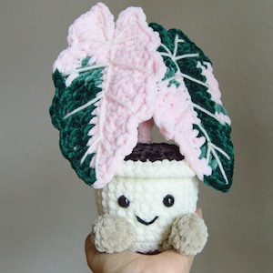 May include: A handmade crochet plant with a smiling face. The plant has large pink and green leaves and is potted in a white pot. The pot has small tan arms. The plant is a cute and whimsical decorative item.