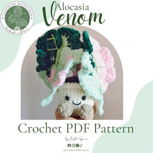 May include: A crochet Alocasia Venom plant with large, variegated leaves in shades of dark green, light green, and soft pink. The plant is potted in a white, textured container featuring a smiling face with black eyes and a stitched mouth.