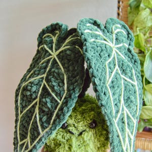 May include: A handmade, plush green plant with large, textured leaves and a smiling face. The leaves have light green veins. The plant is held in a hand, with a background of other plants.