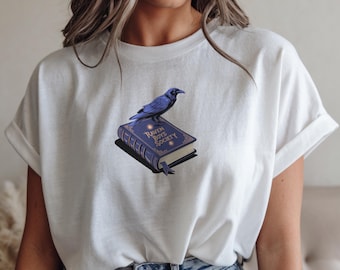 May include: White t-shirt with a graphic of a black raven perched on a closed book. The book is blue with gold accents and the text "Raven Boys Society" on the cover.