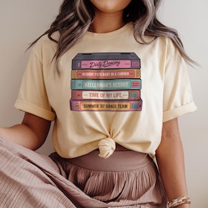 Op de afbeelding: Een lichtgeel T-shirt met een stapel vintage VHS-banden met de film Dirty Dancing. De banden zijn gelabeld met de filmtitel en andere filmgerelateerde tekst, waaronder "Nobody puts baby in a corner", "Kellerman's Resort", "Time of my life" en "Summer '87 Dance Team".