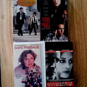 May include: Four VHS movie tapes featuring John Grisham's The Rainmaker, A Time to Kill, Safe Passage, and Conspiracy Theory. Each rectangular case displays film titles, actor names, and promotional imagery including portraits and scenes from the movies.