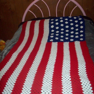 May include: A crocheted blanket resembling the American flag. It has red and white stripes and a blue section with white star shapes. The blanket is displayed on a bed, with a pink metal headboard visible.