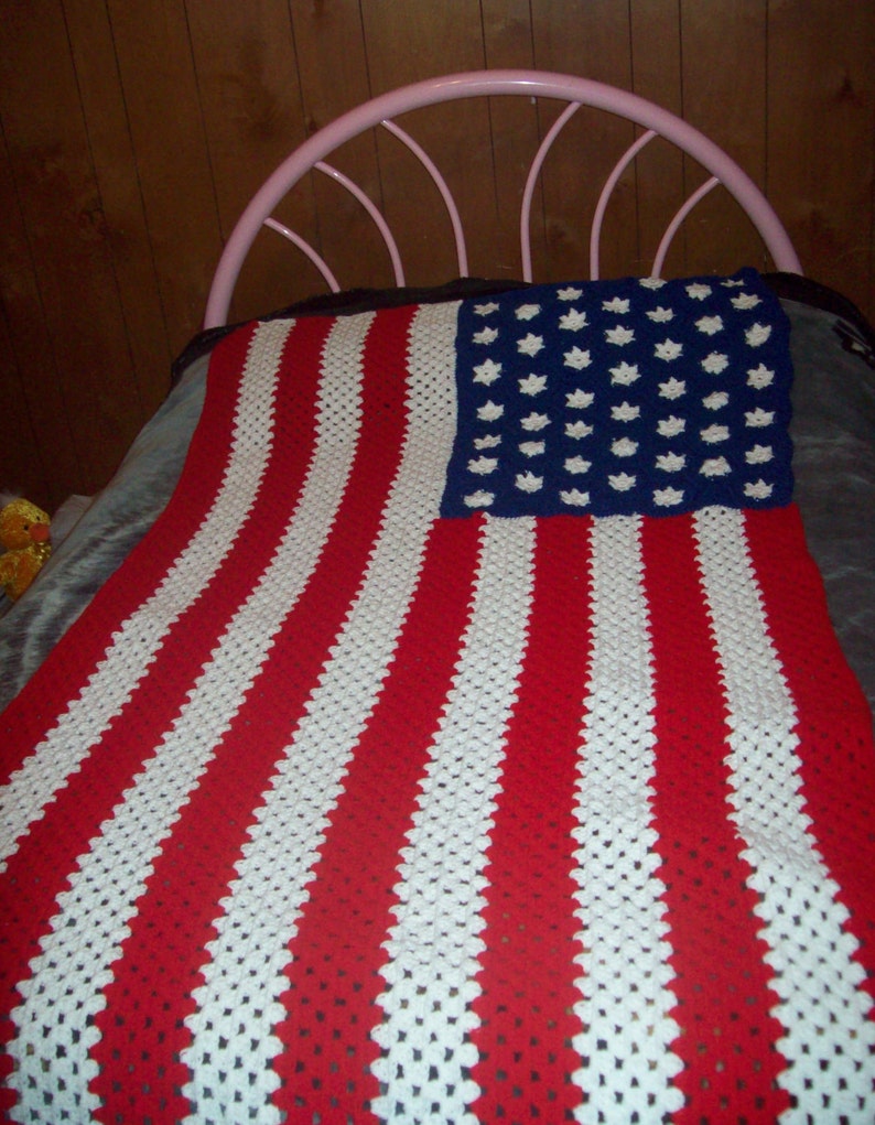 May include: A crocheted blanket resembling the American flag. It has red and white stripes and a blue section with white star shapes. The blanket is displayed on a bed, with a pink metal headboard visible.