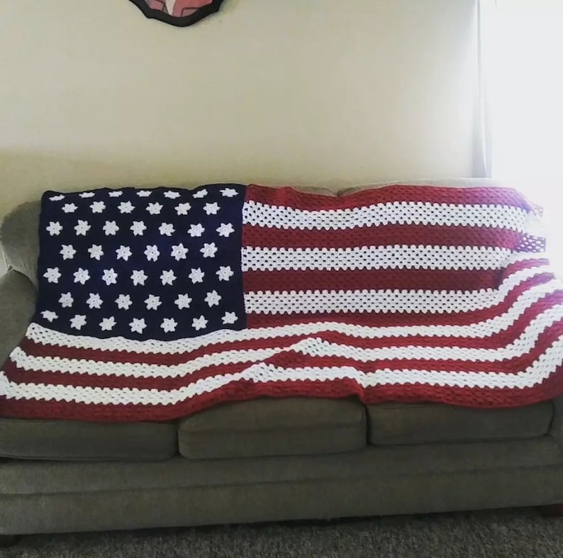 May include: A crocheted blanket in the design of the American flag, draped over a sofa. The blanket features a blue section with white star patterns and red and white stripes. The blanket is made from a soft, textured yarn.