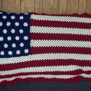 May include: A crocheted blanket in the style of the American flag. It has a blue section with white star-shaped flowers and red and white stripes. The edges are scalloped. The blanket is likely made of wool or a similar yarn.