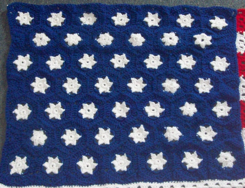 May include: A crocheted blanket in a design inspired by the American flag. The blanket features a blue field with white star motifs, a white border, and a red and white striped section. The blanket is made of yarn and is handmade.