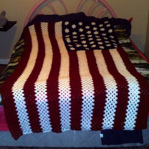 May include: A crocheted blanket resembling the American flag, with red and white stripes and a blue section with white star shapes. The blanket is displayed on a bed, highlighting its handmade quality and patriotic theme.