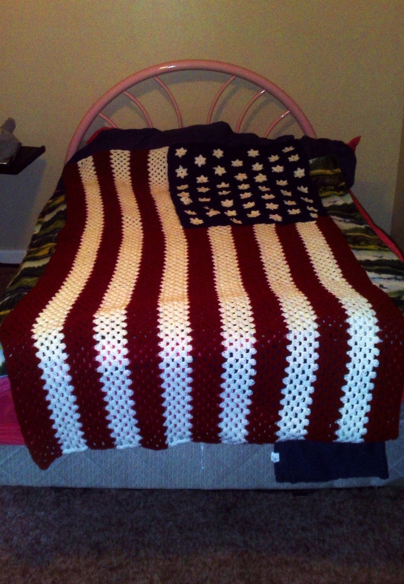 May include: A crocheted blanket resembling the American flag, with red and white stripes and a blue section with white star shapes. The blanket is displayed on a bed, highlighting its handmade quality and patriotic theme.