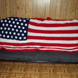 May include: A crocheted blanket in the style of the American flag, draped over a futon. The blanket has a blue section with white star shapes, and red and white stripes. The blanket is made of yarn.