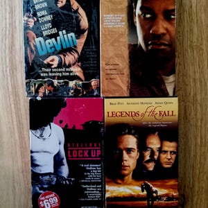 May include: Four VHS movie tapes titled Devlin, John Q., Lock Up, and Legends of the Fall. Each rectangular case features movie poster artwork, cast names, and promotional text. The covers display various photographic scenes and actors.