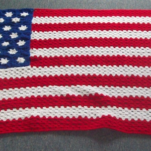 May include: A crocheted blanket depicting the American flag. The blanket has a blue square with white stars in the top left corner and red and white stripes. The blanket is made of yarn and would be suitable for a bed or sofa.