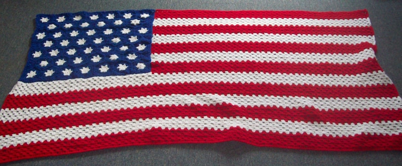 May include: A crocheted blanket depicting the American flag. The blanket has a blue square with white stars in the top left corner and red and white stripes. The blanket is made of yarn and would be suitable for a bed or sofa.