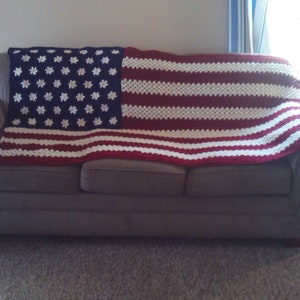 May include: A crocheted blanket in the design of the American flag, draped over a gray sofa. The blanket features a blue section with white star patterns and red and white stripes.