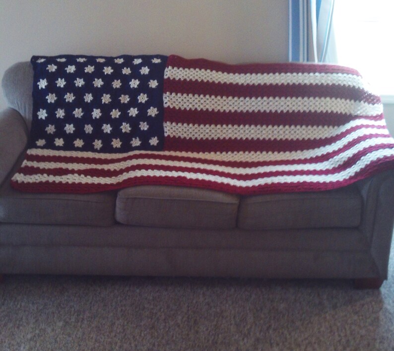 May include: A crocheted blanket in the design of the American flag, draped over a gray sofa. The blanket features a blue section with white star patterns and red and white stripes.