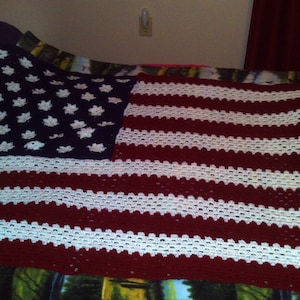 May include: A crocheted blanket in the style of the American flag. It has a navy blue section with white star patterns and red and white stripes. The blanket is handmade and is shown on a bed.