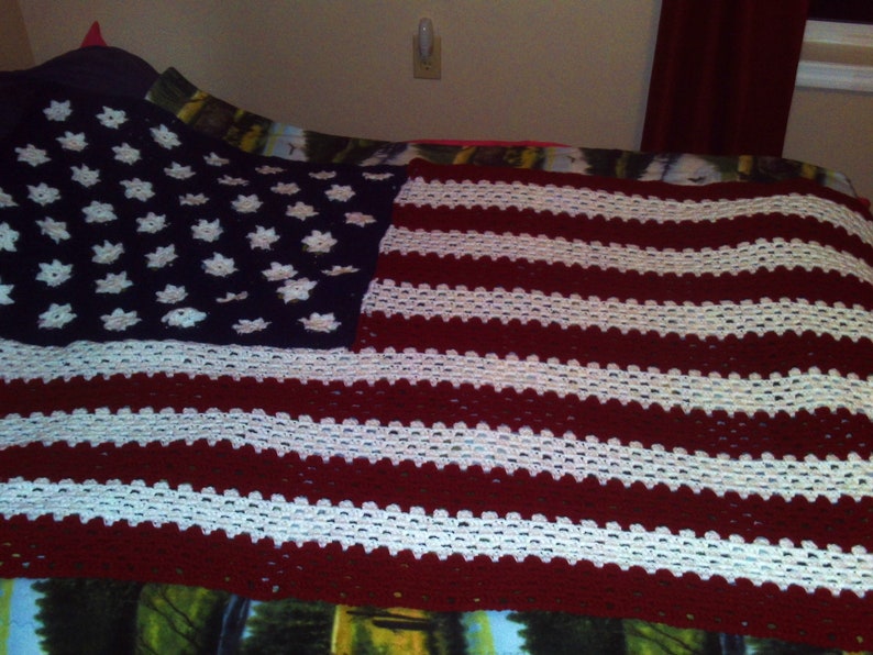 May include: A crocheted blanket in the style of the American flag. It has a navy blue section with white star patterns and red and white stripes. The blanket is handmade and is shown on a bed.
