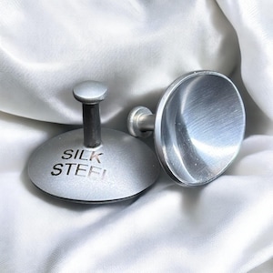 Könnte beinhalten: Zwei silberne Flaschenverschlüsse aus Metall mit runder Oberseite und einem kleinen Stiel. Ein Verschluss hat die Worte "Silk Steel" auf der Oberseite eingraviert.