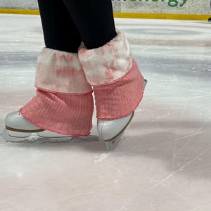 May include: Close-up of ice skates with pink and white leg warmers on an ice rink. The skates are white with silver blades. The leg warmers are pink with a ribbed texture and a white, tie-dye-like trim.