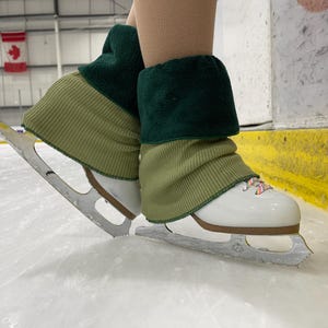 May include: White ice skates with brown soles and silver blades are shown on an ice rink. The skates are paired with green and olive-green leg warmers. The image is taken indoors, with a Canadian flag in the background.