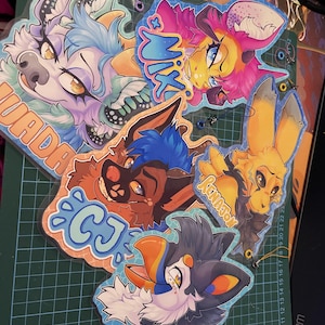 May include: A collection of five colourful acrylic character charms featuring stylised animal illustrations. The charms display the names Nix, CJ, and Renamon in bold lettering, with vibrant artwork showcasing various creatures in shades of blue, pink, yellow, and orange.