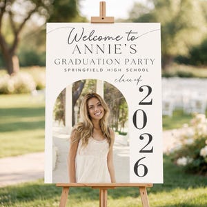 May include: A graduation party welcome sign on a wooden easel. The sign reads "Welcome to ANNIE'S GRADUATION PARTY Springfield High School class of 2026" with a photo of a smiling person.