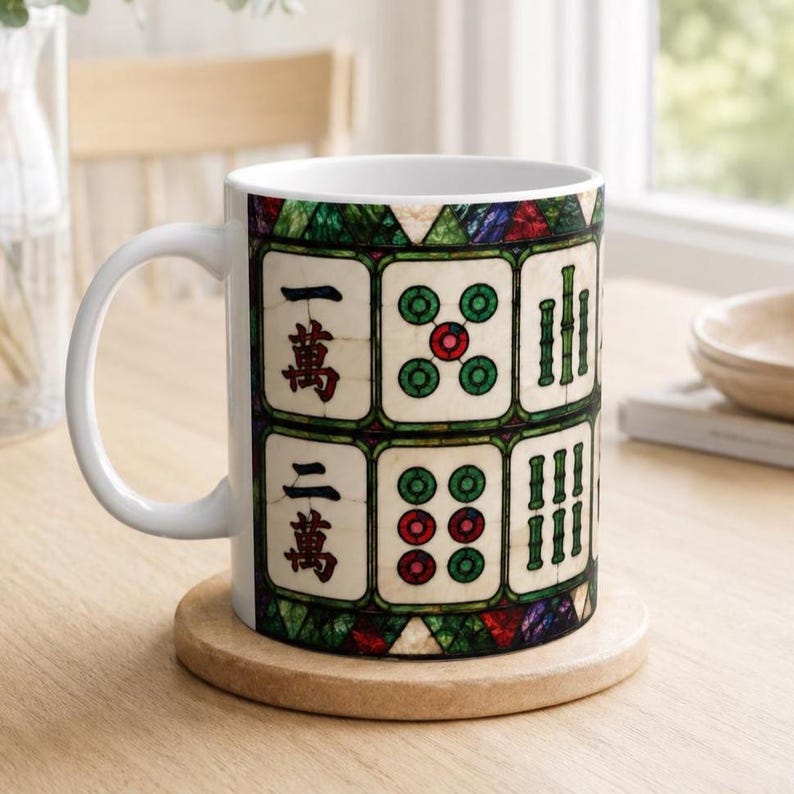 May include: A white ceramic mug featuring a repeating pattern of Mahjong tiles with red and green details. The design includes Chinese characters and bamboo symbols arranged in a grid, framed by a stained glass style border in green and red.