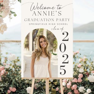 May include: A graduation party welcome sign on a wooden easel. The sign displays the text "Welcome to ANNIE'S GRADUATION PARTY" and "class of 2025". A photo of a smiling young woman is in the centre of the sign.
