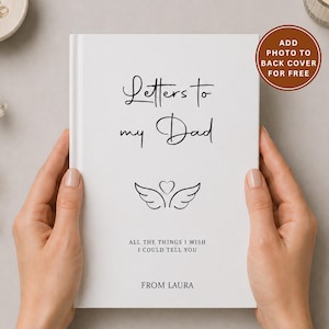 Op de afbeelding: Een wit hardcover dagboek met de titel "Letters to my Dad" in zwart schrift. De omslag heeft een hart- en vleugelontwerp en de tekst "All the things I wish I could tell you" en "From Laura". Een cirkelvormige afbeelding zegt "Voeg gratis een foto toe aan de achterkant".