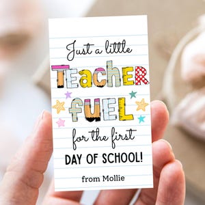 Puede incluir: Una tarjeta blanca con diseño de papel rayado azul, sostenida en una mano, presenta el texto "Just a little TEACHER FUEL for the first DAY OF SCHOOL!" en letras coloridas y juguetonas. La tarjeta está firmada "from Mollie" y decorada con ilustraciones de estrellas.