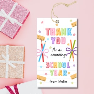 K&ouml;nnte beinhalten: Ein wei&szlig;es Geschenk-Etikett mit dem Text "THANK YOU for an amazing SCHOOL YEAR from Mollie." Das Etikett zeigt farbenfrohen Text, Bleistifte, Lineale und Sterne. Oben ist eine Schnur befestigt. Rosa und goldene Geschenkverpackungen sind im Hintergrund.